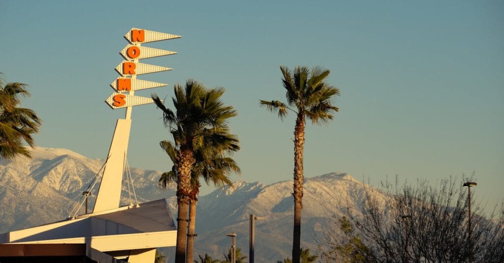A retro Norms restaurant sign framed by palm trees and snow-capped mountains in Rialto, CA.