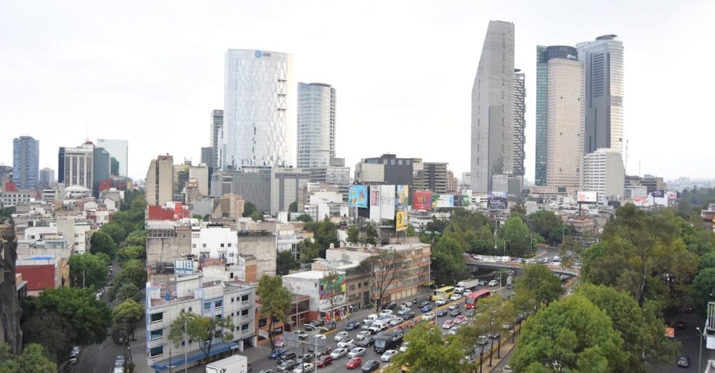 A bustling Mexico City street with skyscrapers, traffic, and greenery under a cloudy sky.