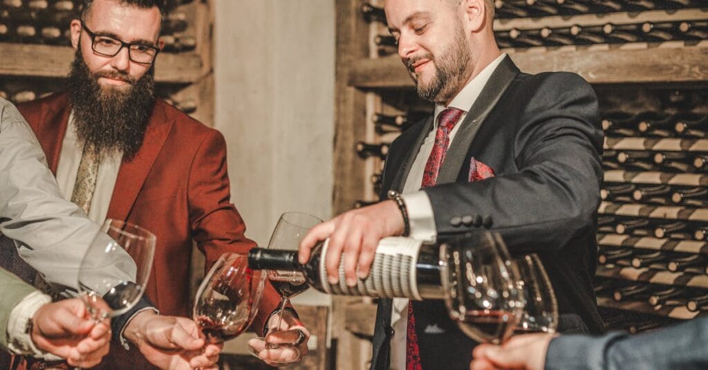 Two men in suits share wine at a stylish cellar gathering.