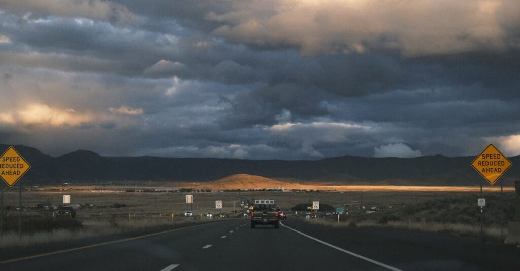 A car drives on a rural highway under dramatic sunset skies, surrounded by mountains.