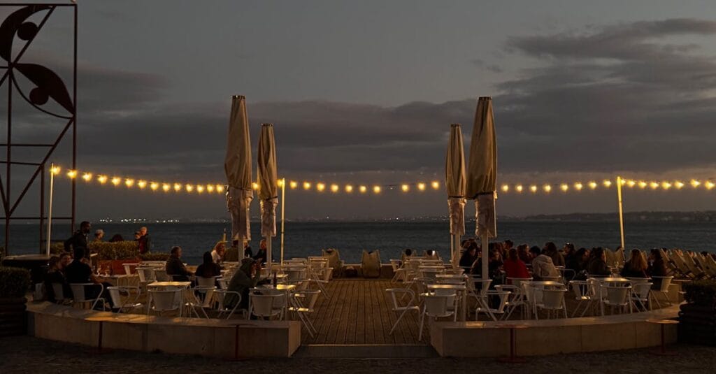 Cozy outdoor dining area by the water at twilight, adorned with glowing string lights.