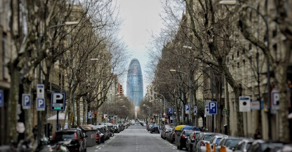 Urban street view of Torre Glòries in Barcelona framed by leafless trees and parked cars.