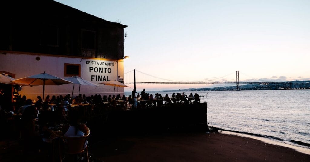 Scenic view of people dining outdoors at Restaurante Ponto Final with Lisbon's sunset and bridge in the background.